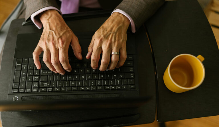 The hands of a senior citizen typing on a keyboard with an emoty coffee mug on the desk next to him.