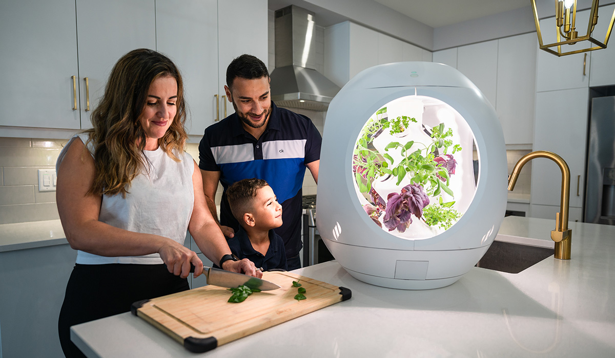 A family of 3 in the kitchen preparing food with the a gardening pod resting on the kitchen counter.