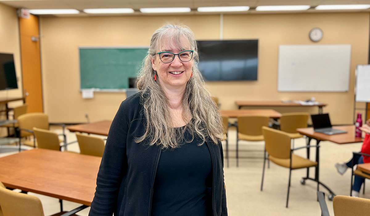 A woman with long gray hair and glasses smiles while posing for a photo in a classroom.