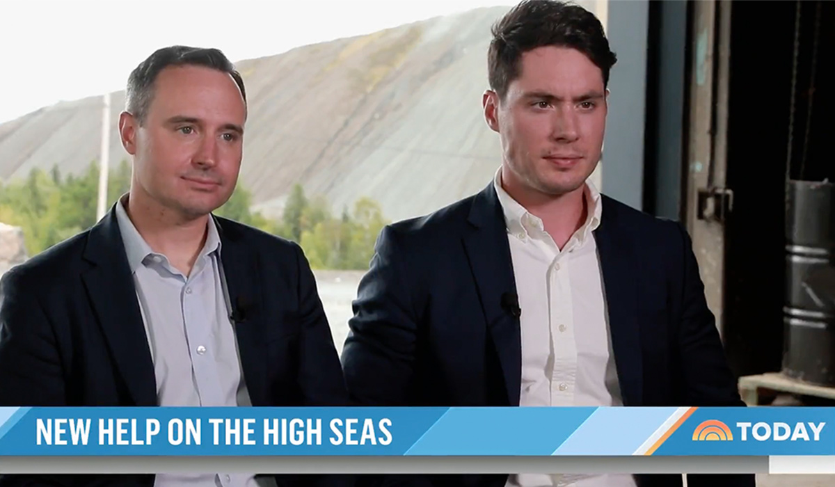 Two men dressed in suits sit for an on camera interview. The words 'New Help on the High Seas' appears on a banner towards the bottom of the screen.