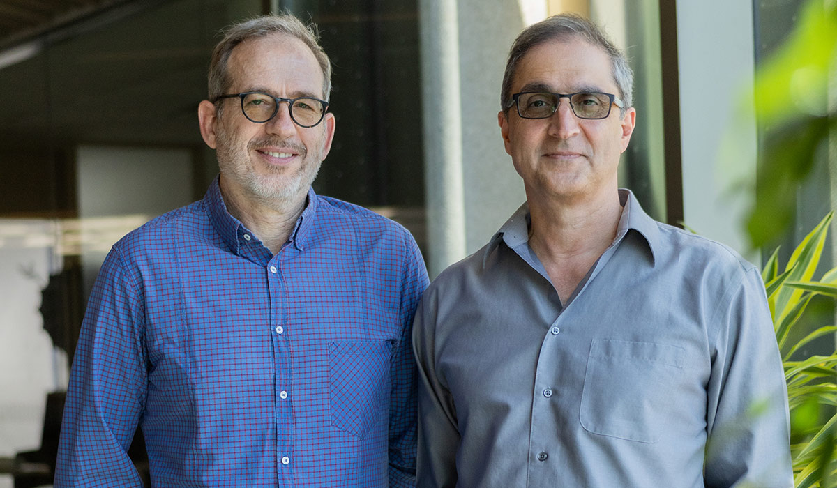 Two older men wearing button up shirts and glasses pose for a photo. 