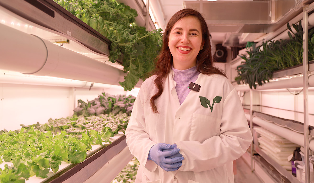A woman in a lab coat smiles for the camera with rows of vegetation visible in the background.