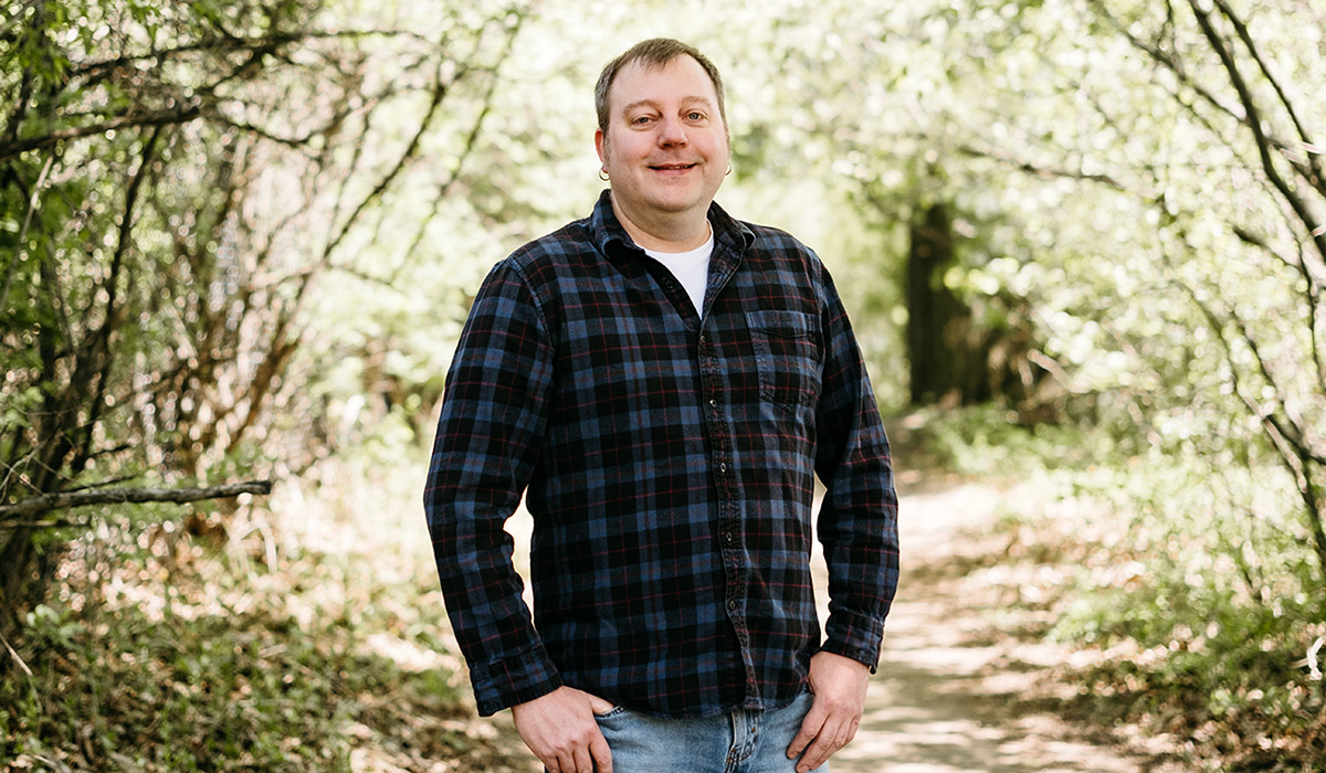 A man in a plaid shirt stands among trees with his thumbs in his pockets.