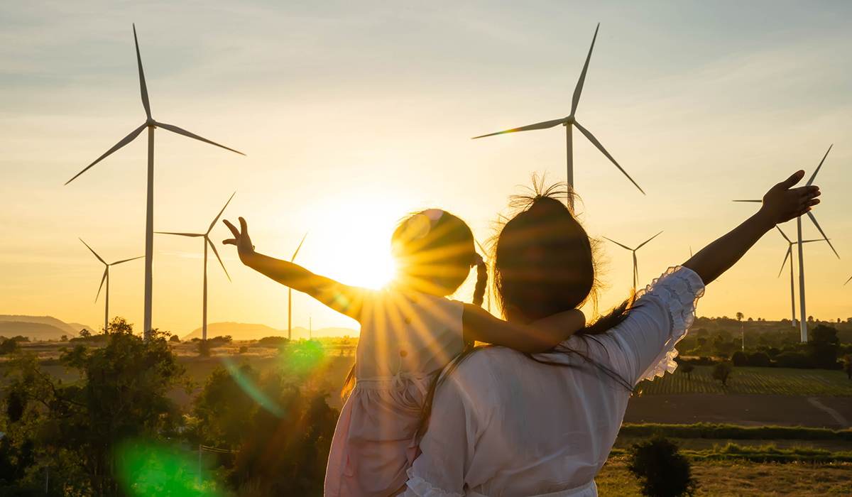 A mother and child spread their arms in celebration with a bright sun and wind turbines visible in the background.