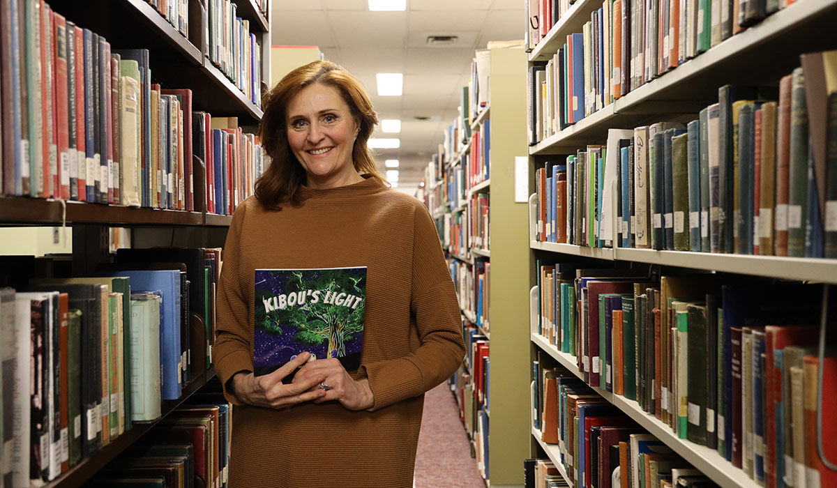 A woman in a brown sweater holds a book in a library.