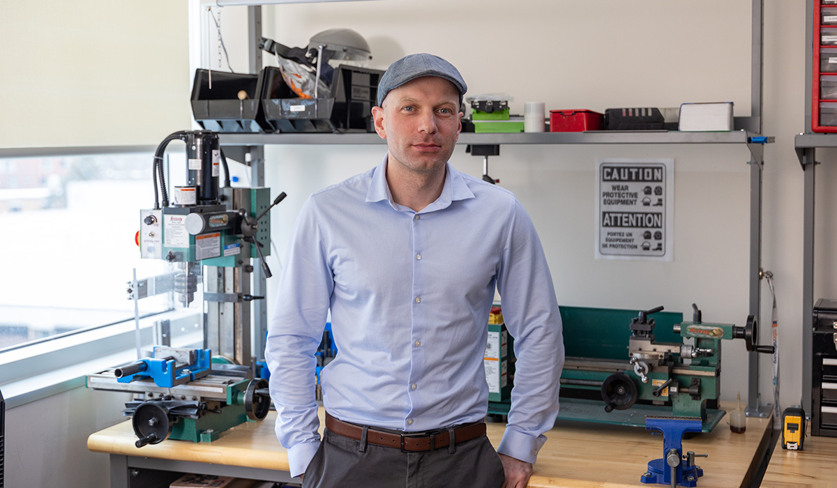 A man in a blue dess shirt and grey hat poses for a photo in lab.