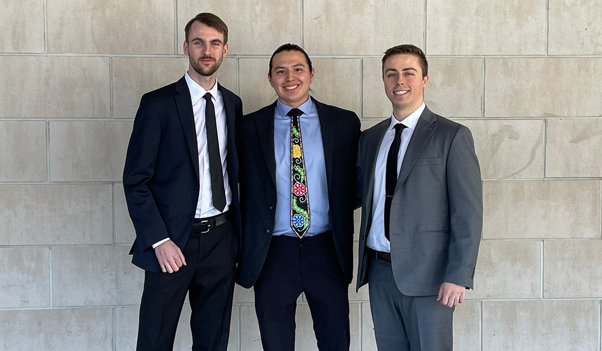 Three men wearing business suits pose for a group photo while standing in front of a stone wall.